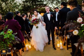In Napa Valley, CA, the happy couple makes their ceremony exit down a candle-lined aisle, surrounded by lush foliage, flowers, and delighted guests, creating a scene of joy and natural beauty.